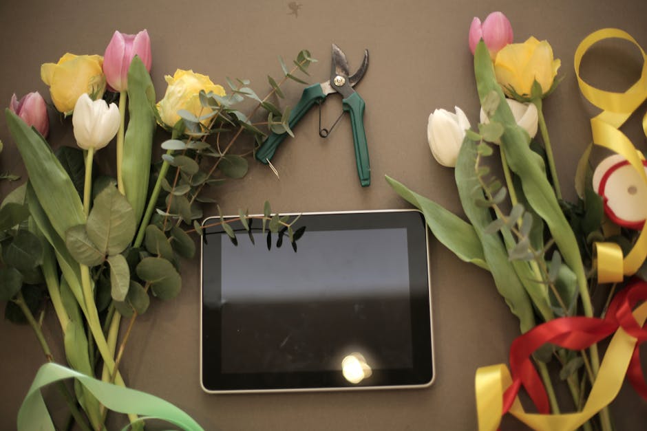 A flat lay arrangement of fresh flowers, including pink and white tulips and yellow roses, with green foliage arranged around a digital tablet. The flowers are in various stages of bloom, with petals 