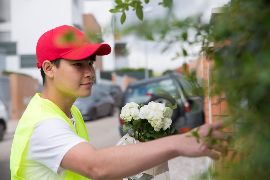 A young male florist wearing a red cap and a yellow high-visibility vest is placing a floral gift of fresh white hydrangeas into a bush or hedge. The arrangement is in a white paper wrapping, and the 