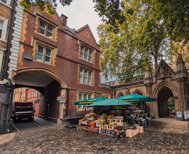 A flower stall situated on a cobblestone street next to an ornate stone archway and historic brick building, featuring a large green umbrella with the website 'floweronwellington.co.uk' printed on it.