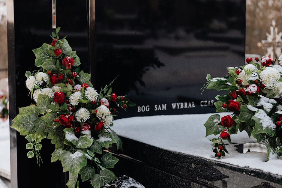 A floral arrangement of red roses, white chrysanthemums, and green ivy leaves placed on either side of a black memorial plaque at a gravesite, with snow scattered across the surface. The flowers are f