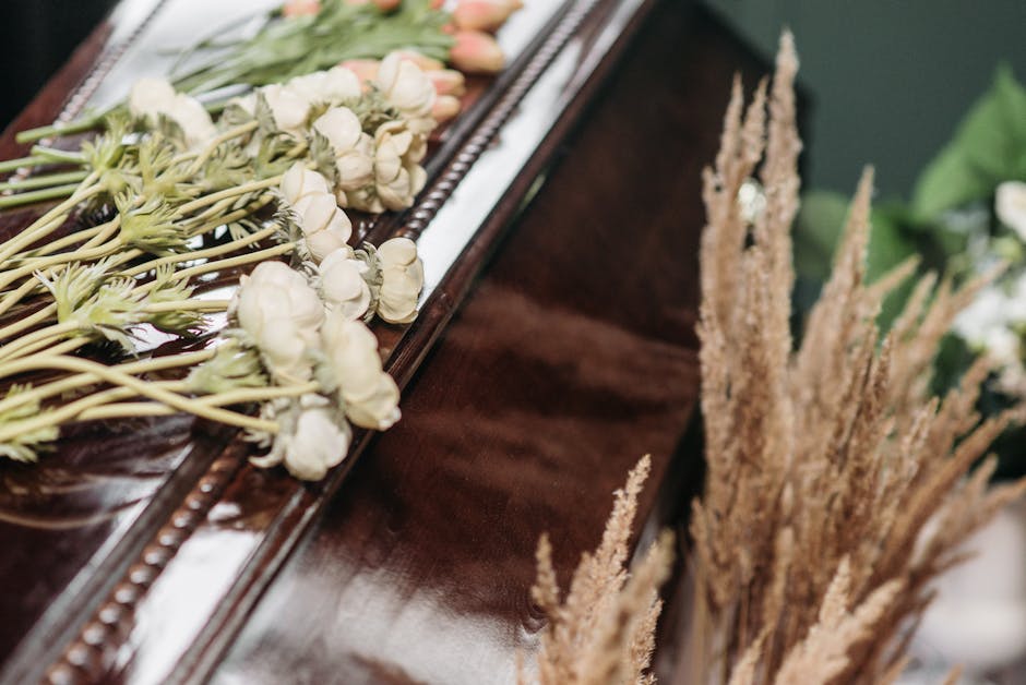 A close-up of a floral arrangement on a polished wooden surface, featuring white and pale pink ranunculus blossoms with tightly packed petals and green stems, alongside beige dried pampas grass with f