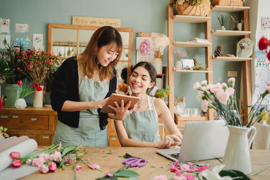 A florist and a customer in a flower shop engaging in a conversation while looking at a tablet. The florist, wearing a light green apron over a black cardigan, is smiling and holding the tablet, showi