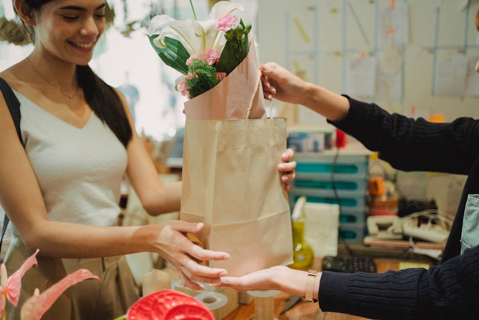 A woman with long dark hair, dressed in a white sleeveless top, is smiling as she receives a bouquet of fresh flowers from a florist in a workshop. The bouquet, wrapped in pink paper, contains white l